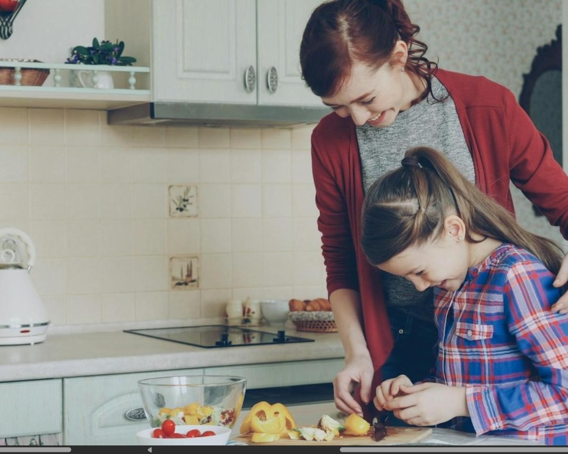 A mother cooking at the kitchen counter in warm afternoon light while her daughter sits nearby at the table, focused on a drawing